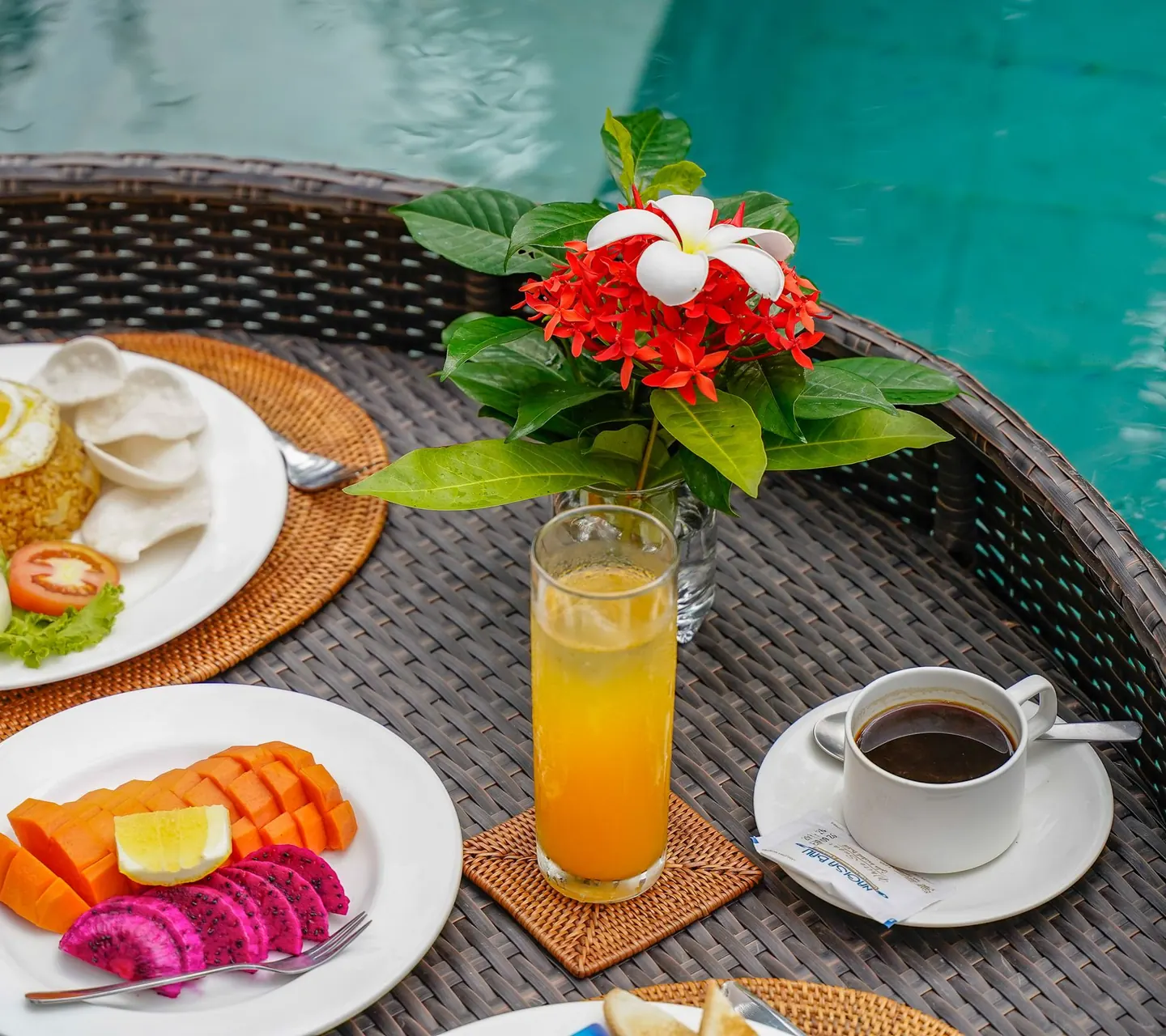 A rattan tray floating in the pool with fruit, orange juice, coffee and a sprig of flowers