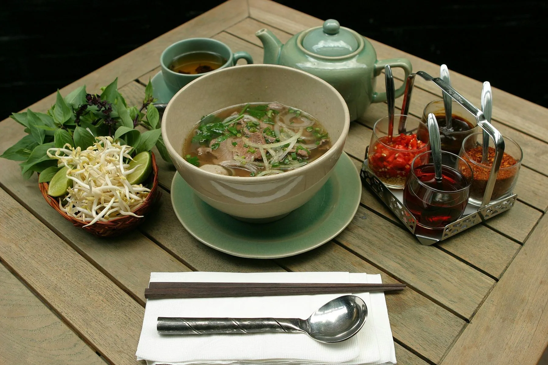 A bowl of phở on a wooden table in Hoi An, with fresh herbs, bean sprouts, lime and the chilli and sauce tray