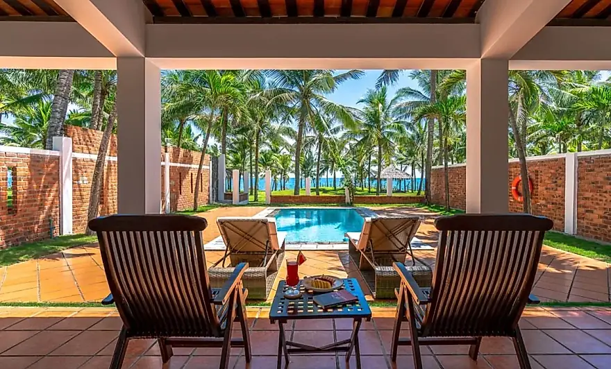 Terraza cubierta de la villa con dos butacas y una mesa, la piscina privada delante y el mar tras las palmeras
